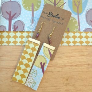 Larger rectangle dangle scrapbook paper earrings with gold metal accents and purple/blue irridescent beads. Front is light blue with trees and back is gold/yellow fan shaped. Front and back are shown. Earrings are displayed on wooden table with corresponding scrapbook paper behind them.