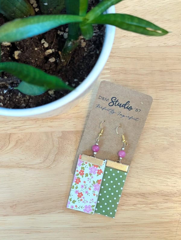 Floral earrings green, orange, and pink flowers with a cream background on the front side are shown. The back side is green with white polkadots. Pink beads and gold metals. Earrings are displayed on a wooden table with a plant next to it.