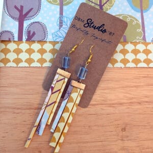 Thin triple high-low rectangle scrapbook earrings with gold metal accents and blue beads. Gold and yellow with blue, purple, and green trees. Front and back are shown. Earrings are displayed on wooden table with corresponding scrapbook paper behind them.