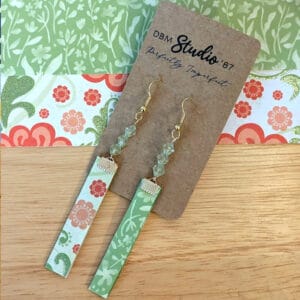 Floral earrings with green, orange and red flowers on the front, green and cream flowers on the back. Green beads and gold metals. Earrings are displayed on a wooden table with corresponding scrapbook paper below it.