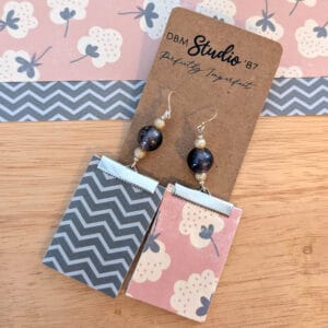 Medium rectangle dangle scrapbook paper earrings with silver metal accents with silver and purple pearlish beads. Front is a salmon pink with white and gray cotton looking flowers. Back is dark and light gray chevron. Front and back are shown. Earrings are displayed on wooden table with corresponding scrapbook paper behind them.