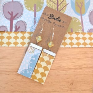 Medium rectangle dangle scrapbook paper earrings with silver metal accents and yellow clear crytsal shaped beads. Front is light blue with trees and back is gold/yellow fan shaped. Lightweight Mod Podged scrapbook paper with felt core. Front and back are shown. Earrings are displayed on wooden table with corresponding scrapbook paper behind them.