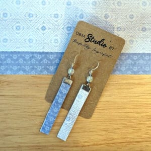 Thin rectangle dangle scrapbook paper earrings with silver metal accents and blue sparkly beads. Light blue floral and linear front and darker blue floral back. Front and back are shown. Earrings are displayed on wooden table with corresponding scrapbook paper behind them.