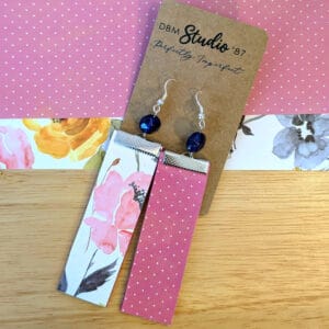 Floral earrings with Pinkish assorted flowers on the front, pink and white polka dots on the back. Blue beads and silver metal hooks. Earrings are displayed on a wooden table with corresponding scrapbook paper below it.