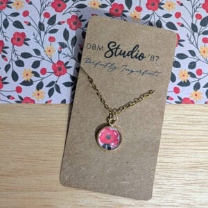 A dainty gold chain necklace with a small glass cabochon pendant featuring a red flower on a soft pink background, displayed on a wooden table with matching floral paper beneath it.
