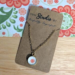 A dainty gold chain necklace with a small glass cabochon pendant featuring a reddish-orange flower on a off-white background, displayed on a wooden table with matching floral paper beneath it.