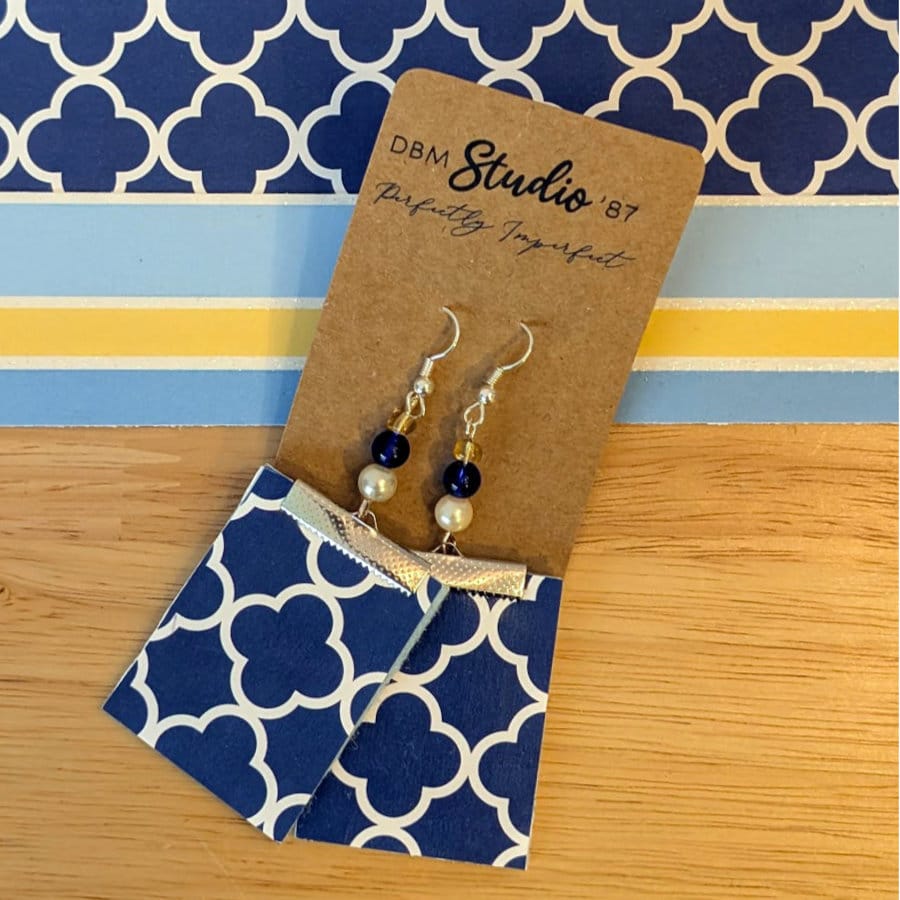 Blue Square Paper and felt earrings in silver tone with blue and silver beads. Back side is shown, dark blue and white clover-ish design. Earrings are on a wooden table with corresponding scrapbook paper shown.