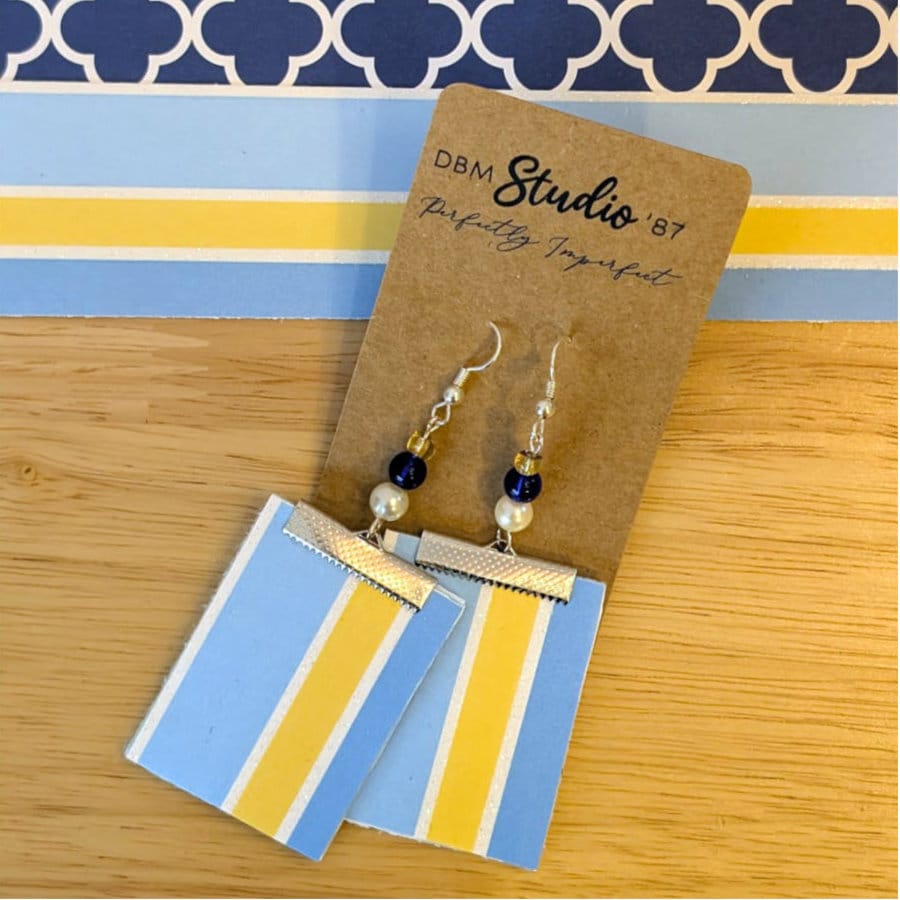 Blue Square Paper and felt earrings in silver tone with blue and silver beads. Front side is shown light blue, yellow and white stripes. Laid on a wooden table with matching scrapbook paper in the background.