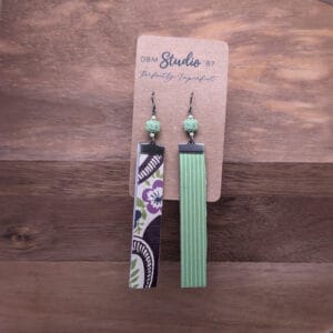 long scrapbook rectangle earrings with florals and swirls of purple, blue, green and brown on the front and green with white vertical stripes on the back. Pewter colored finishes and green and cream beads. Modpodged and sandwhiched with felt. Front and back are shown.