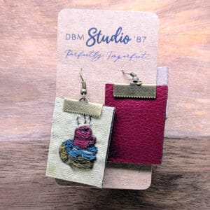 A pair of embroidered earrings with a coffee cup on a stack of books pictured on them. With a maroon back.